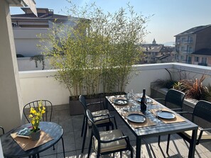 En takterrass med ett matbord för två personer, omgiven av stolar, en krukväxt och en utsikt över byggnader i bakgrunden.