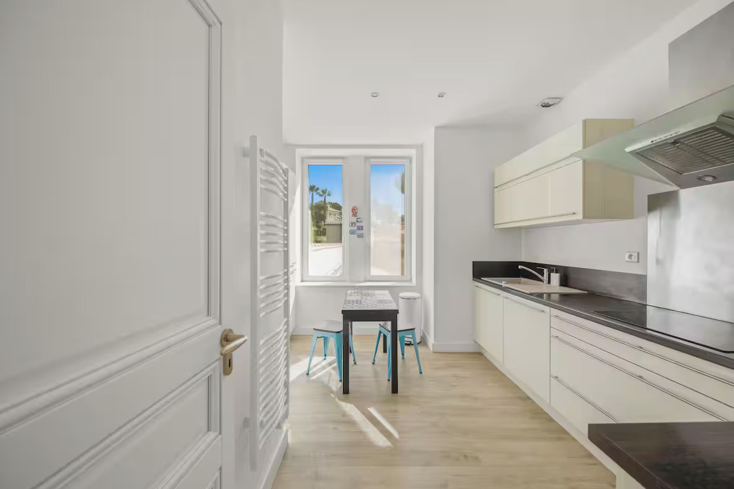 A modern kitchen with white cabinetry, a stainless steel range hood, and a window with a view of palm trees.