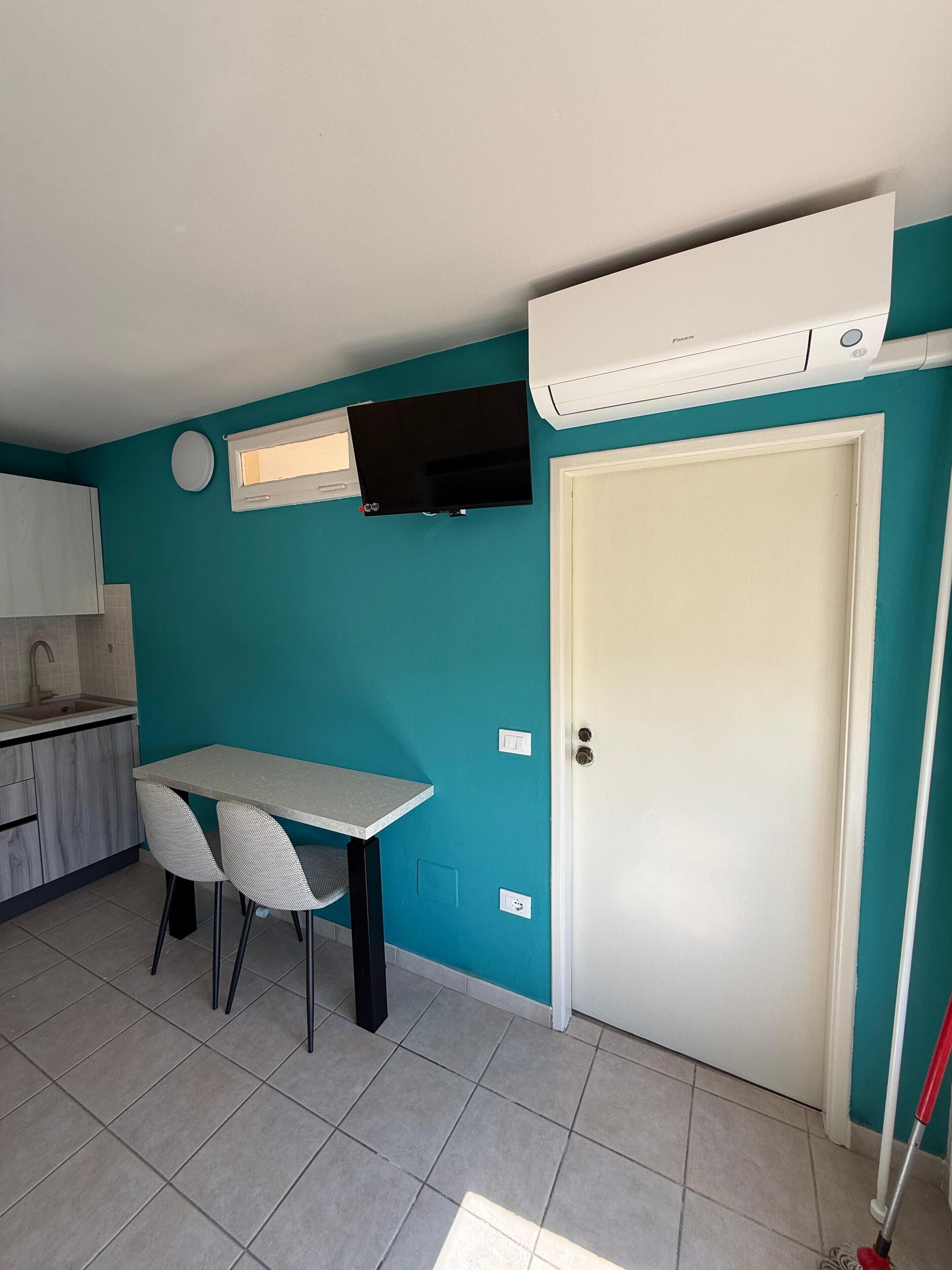 A kitchen with teal walls, a white door, a table with chairs, a TV mounted above the counter, and an air conditioning unit.