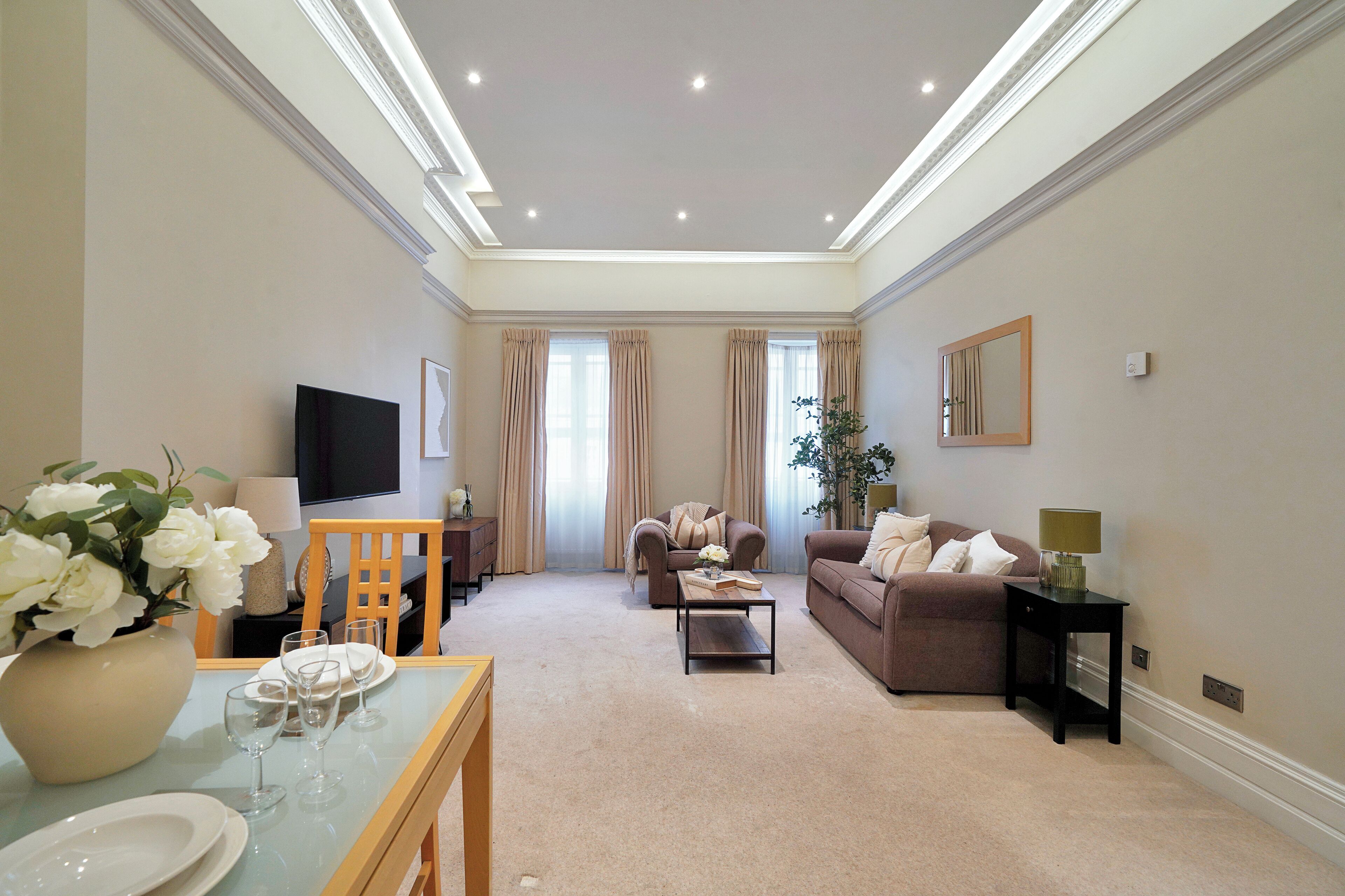 A spacious living room with a flat-screen TV, beige sofas, a dining table set with plates and glasses, and a large vase of white flowers.