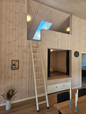 A loft-style room with a wooden interior, a ladder to the upper level, a bed, a clock, and a small shelf with a flag. - New and modern holiday home with hot tub (Grenaa)
