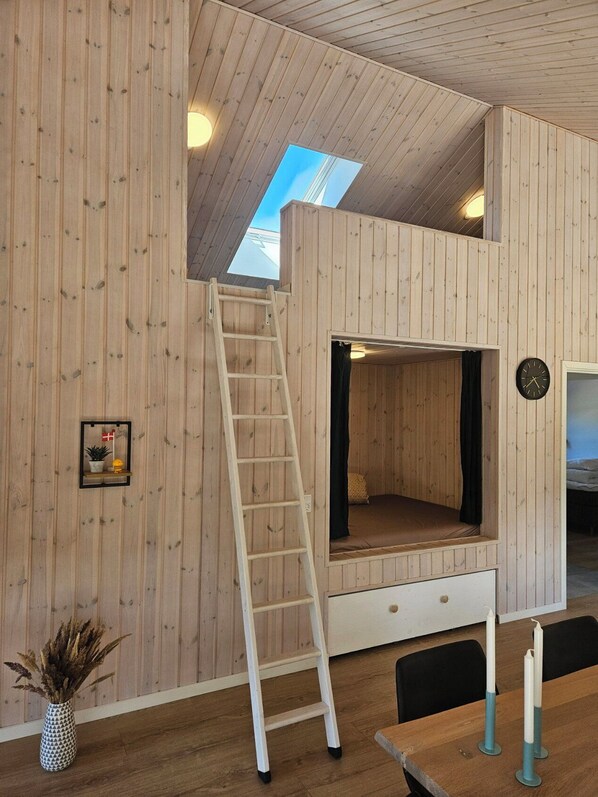 A loft-style room with a wooden interior, a ladder to the upper level, a bed, a clock, and a small shelf with a flag.
