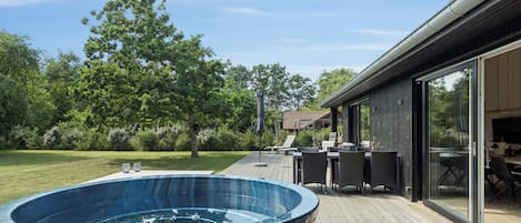 A hot tub on a wooden deck with outdoor furniture and a glass door leading to a covered area.