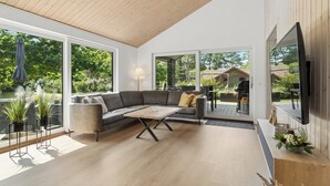 A modern living room with a sectional sofa, a coffee table, and large windows offering a view of the outdoors. - New and modern holiday home with hot tub (Grenaa)