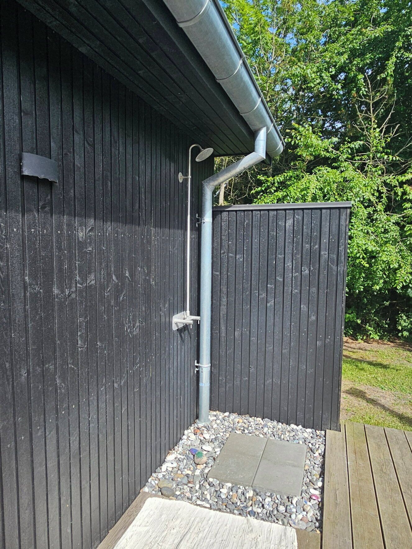 A wooden structure with a metal gutter, a showerhead, and a small outdoor shower area with pebbles and a wooden deck.