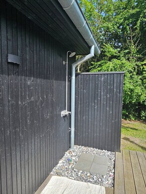 A wooden structure with a metal gutter, a showerhead, and a small outdoor shower area with pebbles and a wooden deck.