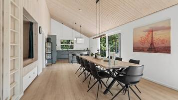 Un comedor espacioso con una larga mesa de madera y sillas modernas, una cocina al fondo y un ventanal con vistas a zonas verdes.