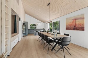 Un vaste espace à manger avec une longue table en bois et des chaises modernes, une cuisine en arrière-plan et une grande fenêtre donnant sur un paysage verdoyant.