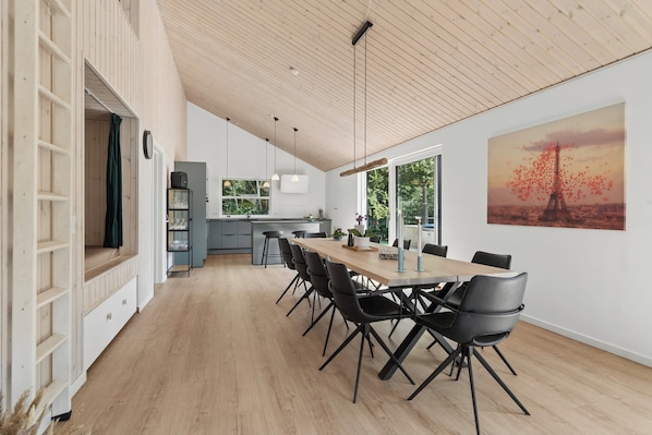 A spacious dining area with a long wooden table and modern chairs, a kitchen in the background, and a large window with a view of greenery. - New and modern holiday home with hot tub (Grenaa)