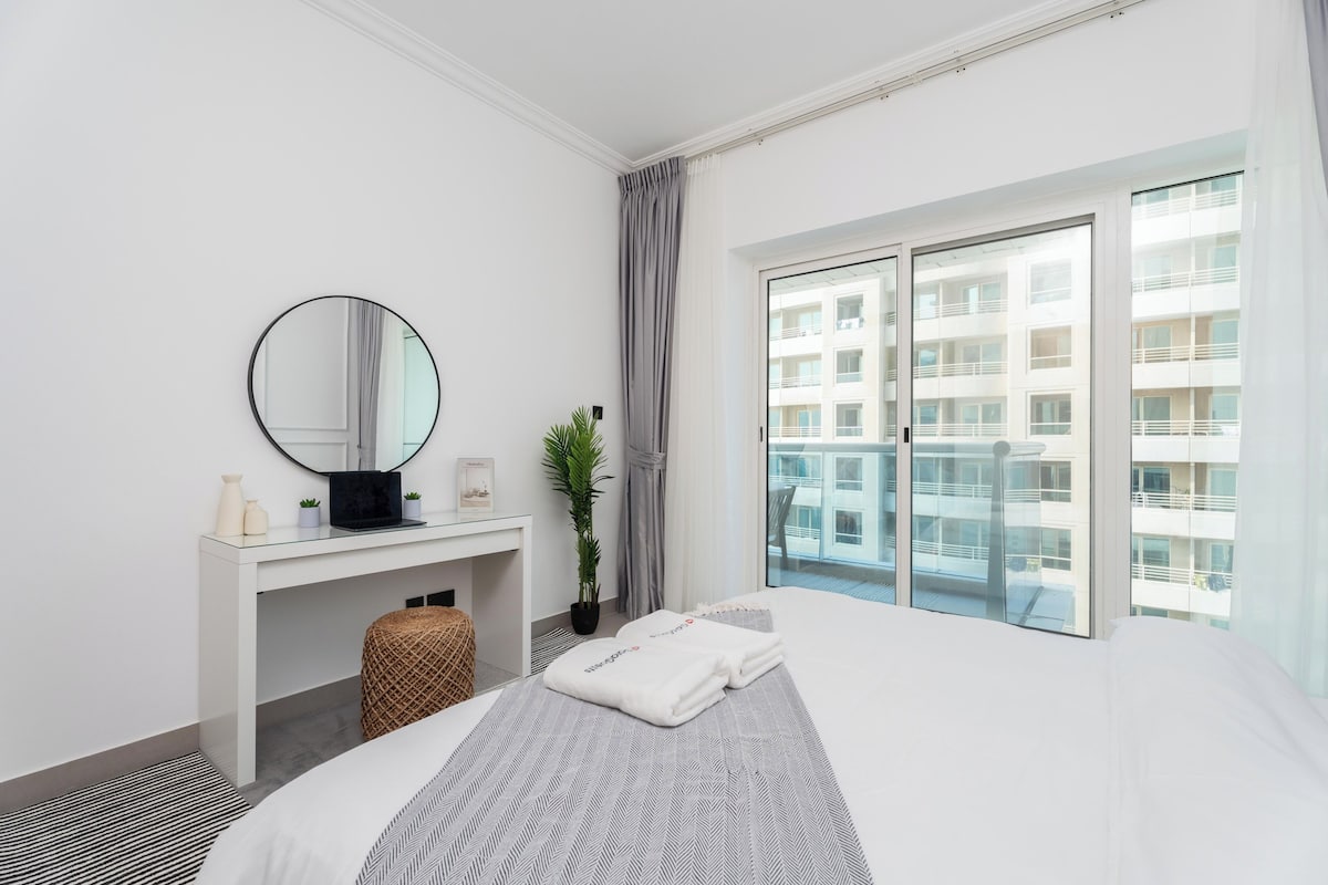 A hotel room with a bed, a desk with a computer, a mirror, a window with a view of buildings, and a potted plant.