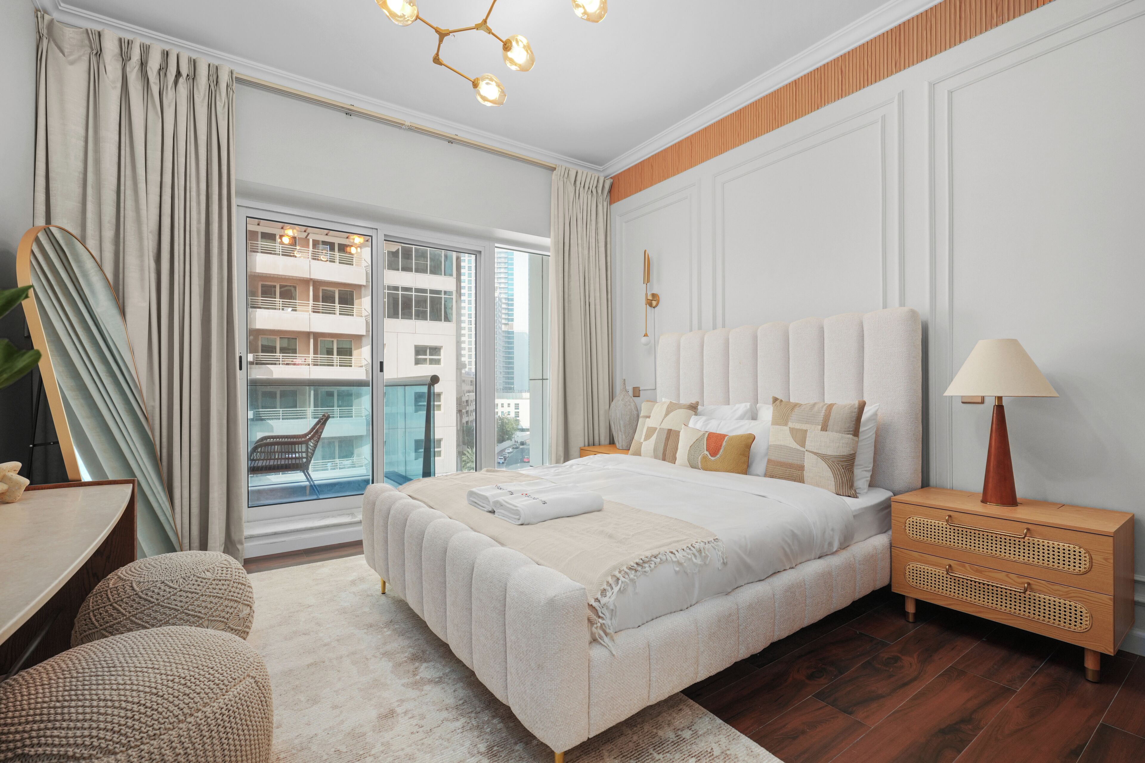 A modern hotel room with a large bed, a wooden nightstand, a desk, and a view of a building through a glass door.