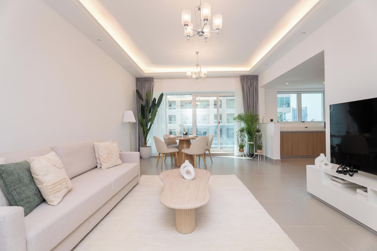 A modern living room with a sofa, a coffee table, a dining table with chairs, a TV, and a balcony with a view.