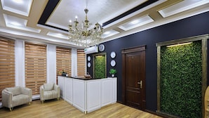 Un’area reception con pavimento in legno, una scrivania, due poltrone, un lampadario e un pannello decorativo con motivi a foglia verde.