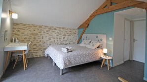 Een slaapkamer met een stenen muur, een bed met beddengoed met een patroon, een bureau met een stoel, en een houten plafondbalk.