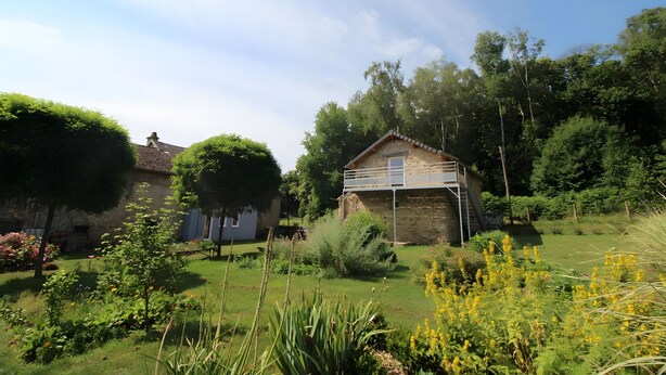 Una casa de piedra con terraza de madera, rodeada de un jardín exuberante con diversas plantas y árboles.