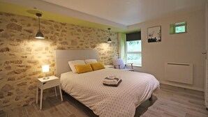 Un dormitorio con pared de piedra, cama con ropa de cama estampada, mesita de noche con lámpara, una silla y ventana con vistas.