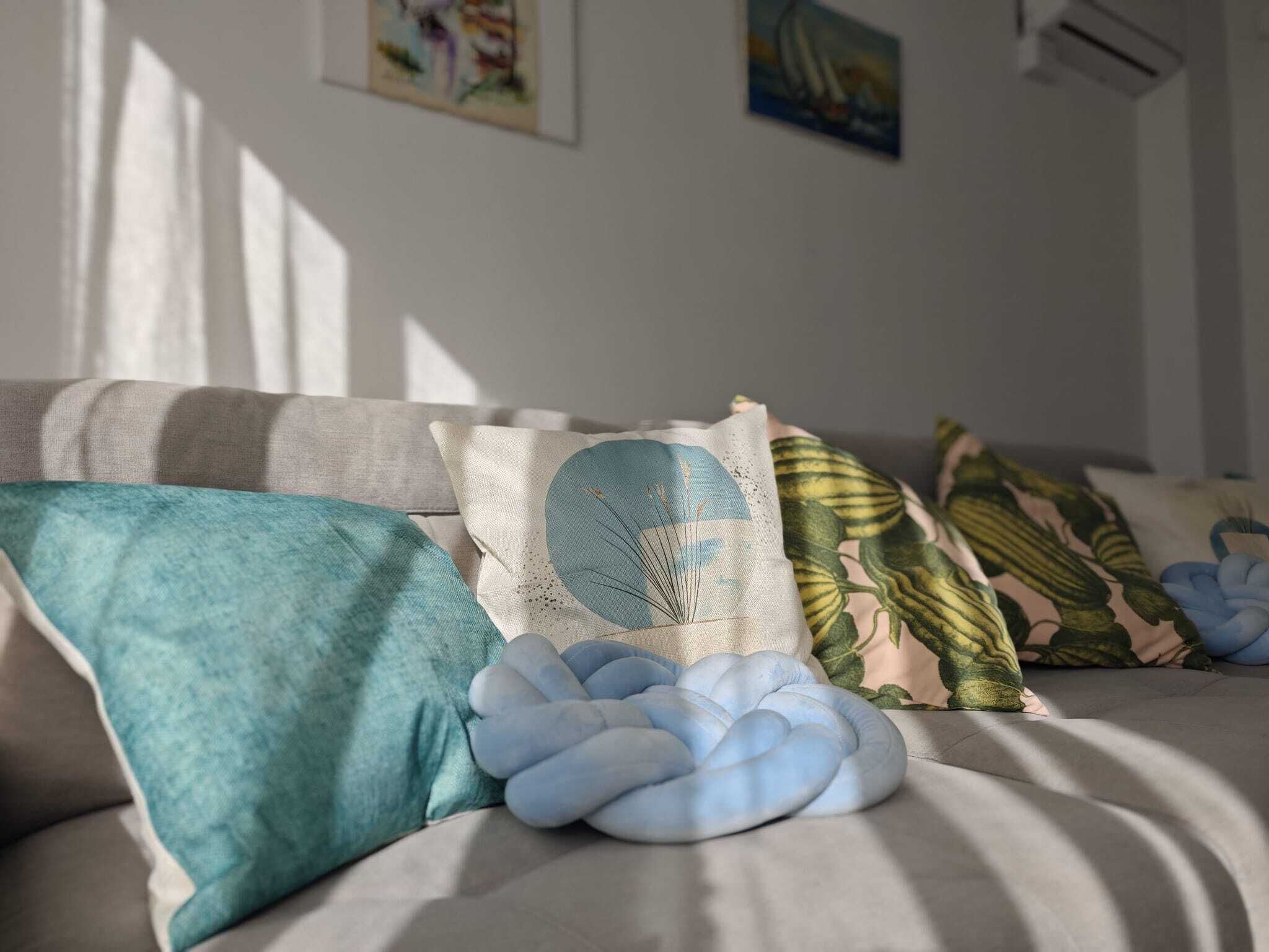 Un sofá con varios cojines, incluyendo uno con forma de nube azul, en una habitación con luz solar entrando por la ventana.