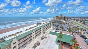 Un hotel con vista a la playa, varios pisos y un estacionamiento con coches y palmeras.