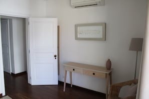 A room with a white door, a wooden console table with a vase, a framed picture on the wall, and a wicker chair. - Apartment 'Cascais Bay Terrace V' with Sea View, Wi-Fi and Air Conditioning (Cascais)