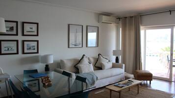 A modern living room with a glass dining table, a sofa with decorative pillows, and a view through a large sliding glass door.