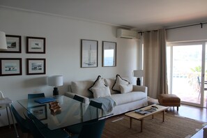 A modern living room with a glass dining table, a sofa with decorative pillows, and a view through a large sliding glass door. - Apartment 'Cascais Bay Terrace V' with Sea View, Wi-Fi and Air Conditioning (Cascais)
