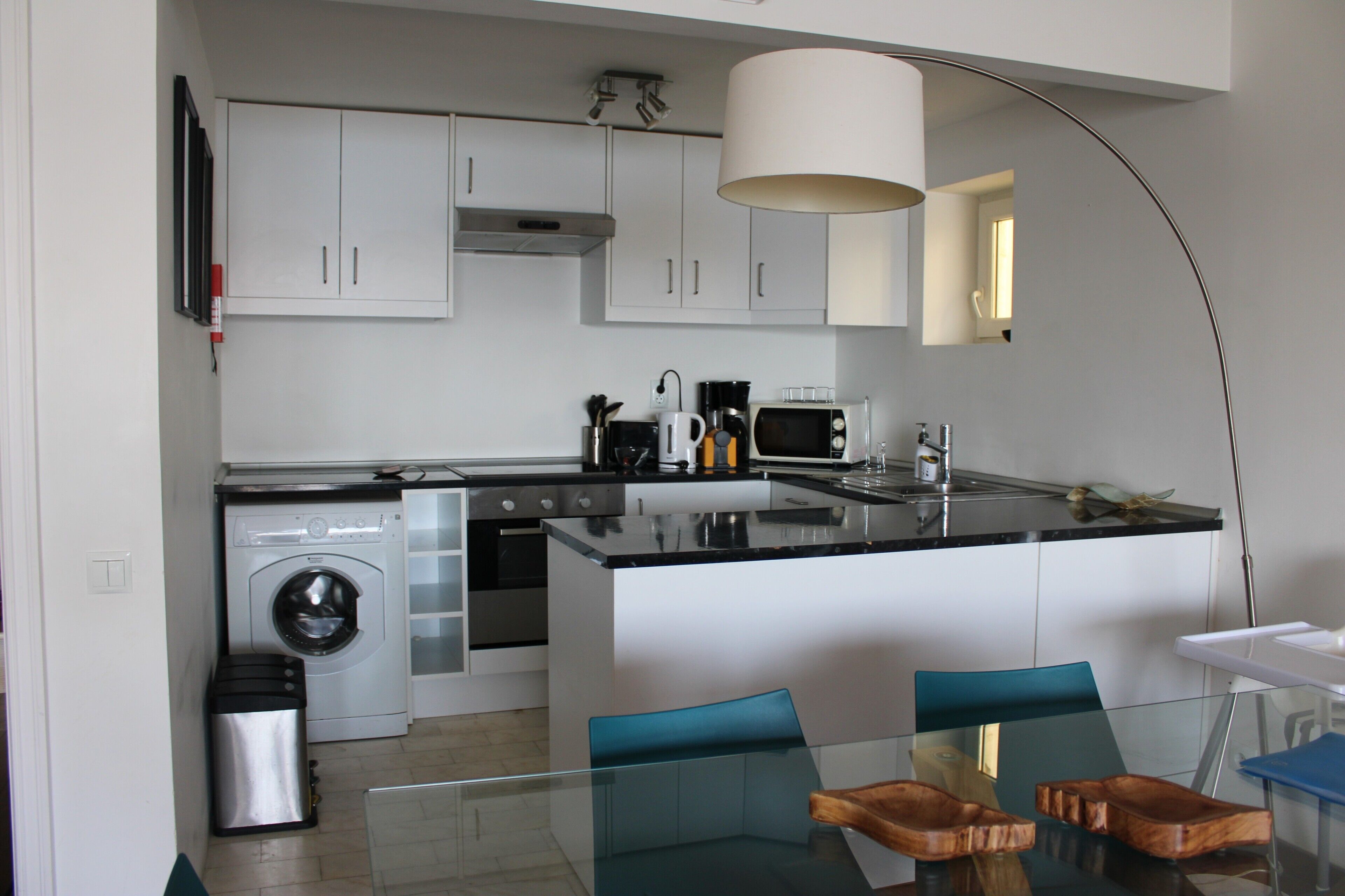 Een moderne keuken met witte kasten, een wasmachine, een magnetron en een glazen eettafel met stoelen.