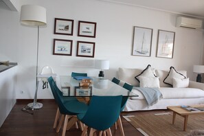 Een moderne woonkamer met een glazen eettafel, blauwgroene stoelen, een witte bank met decoratieve kussens, en kunstwerken in lijsten aan de muur.
