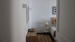A narrow hallway with a wooden bench, a small round table with a lamp, a bed with a white headboard, and a framed picture on the wall. - Apartment 'Cascais Bay Terrace V' with Sea View, Wi-Fi and Air Conditioning (Cascais)