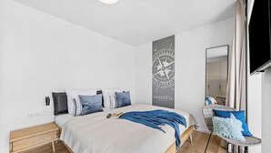 A modern bedroom with a large bed, a wall-mounted TV, and a wooden nightstand. - The View B16 Meeresleuchten - Balcony, Sofa bed, shared Sauna, Elevator (Dagebüll)