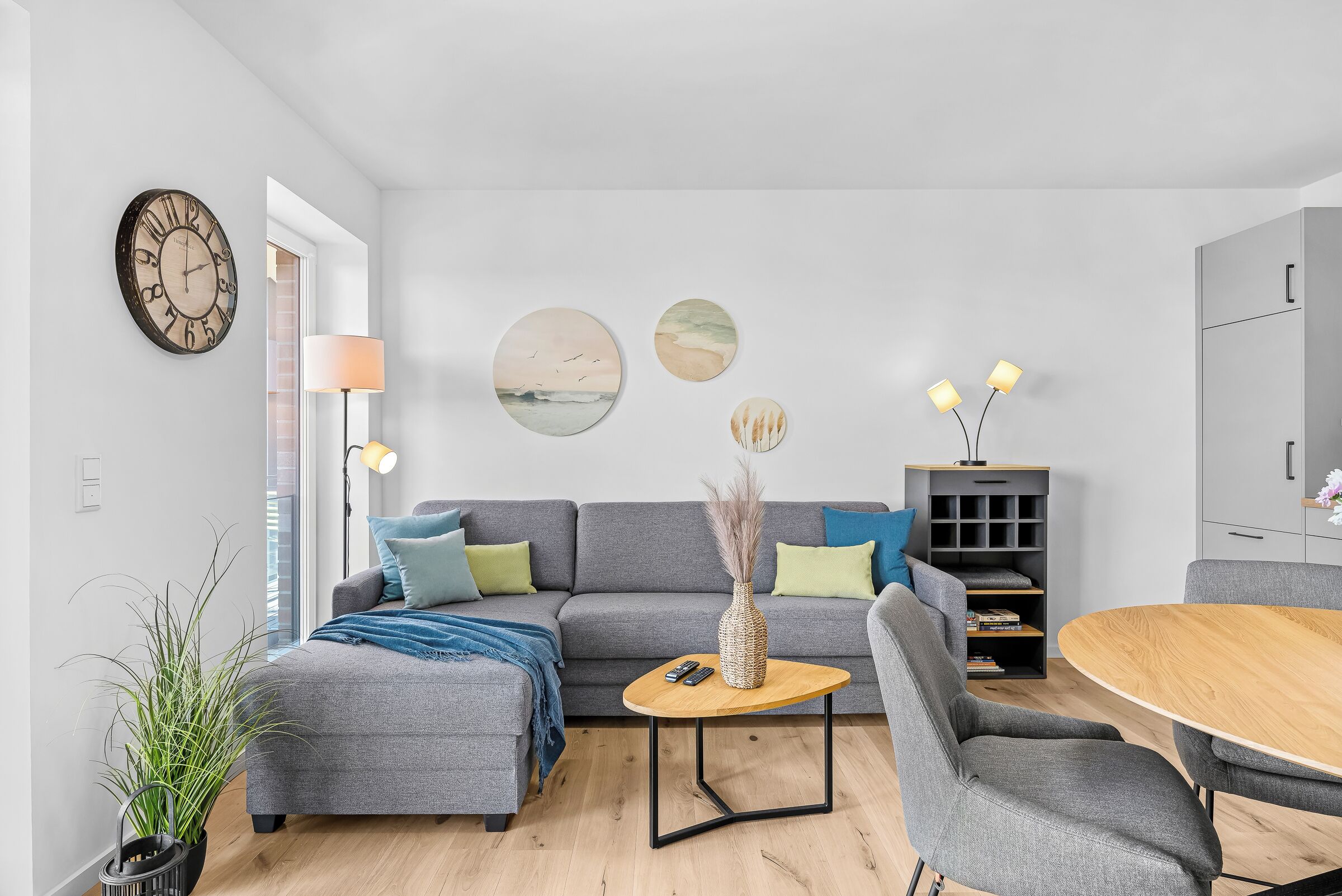 Un salon moderne avec un canapé gris, une table basse et une table à manger. Il y a une horloge au mur et une lampe sur une table d’appoint.