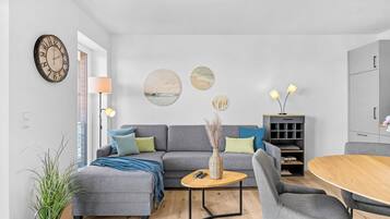 Un salon moderne avec un canapé gris, une table basse et une table à manger. Il y a une horloge au mur et une lampe sur une table d’appoint.