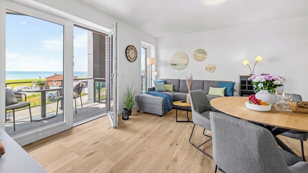 A modern living room with a grey sofa, a wooden dining table, and a balcony with a view of the sea. - The View B16 Meeresleuchten - Balcony, Sofa bed, shared Sauna, Elevator (Dagebüll)