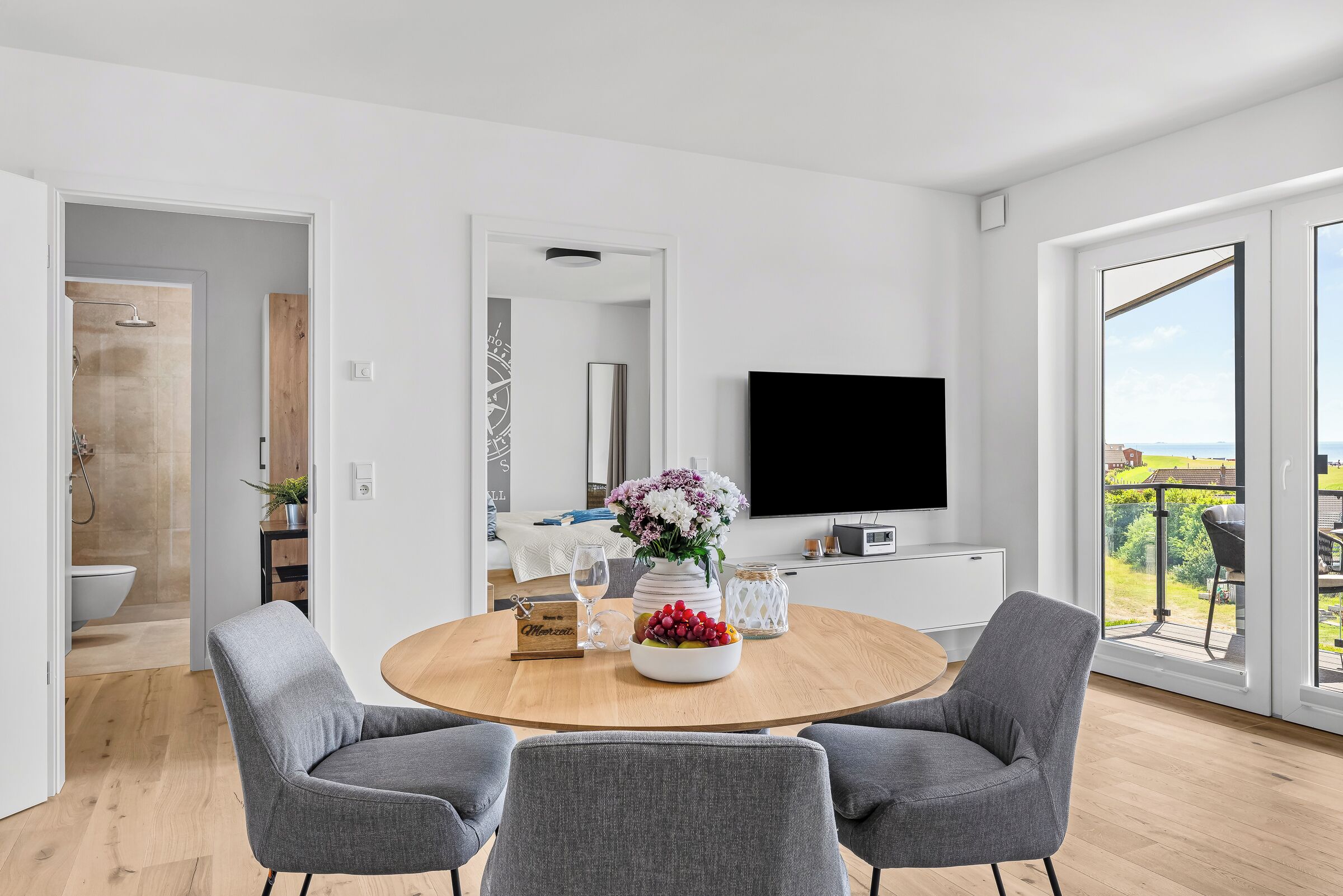 Un salon moderne avec une table à manger ronde en bois, des chaises à revêtement gris, un téléviseur à écran plat fixé au mur et une grande fenêtre donnant sur l’extérieur.
