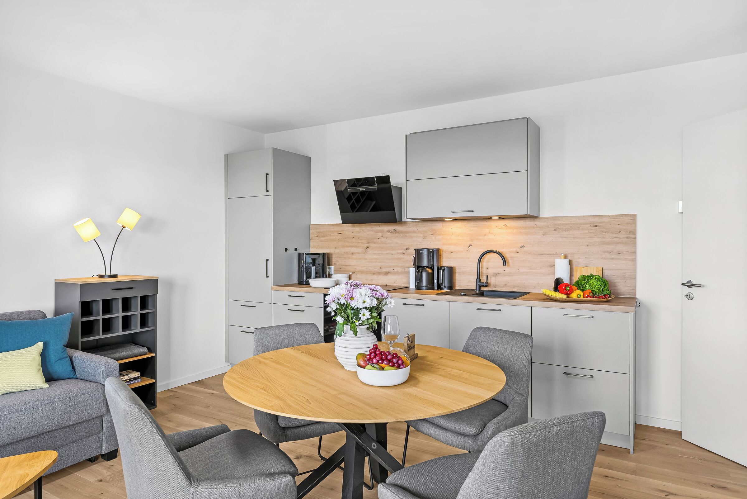A modern kitchen with a dining area, grey furniture, and a wooden table with a fruit bowl and flowers.