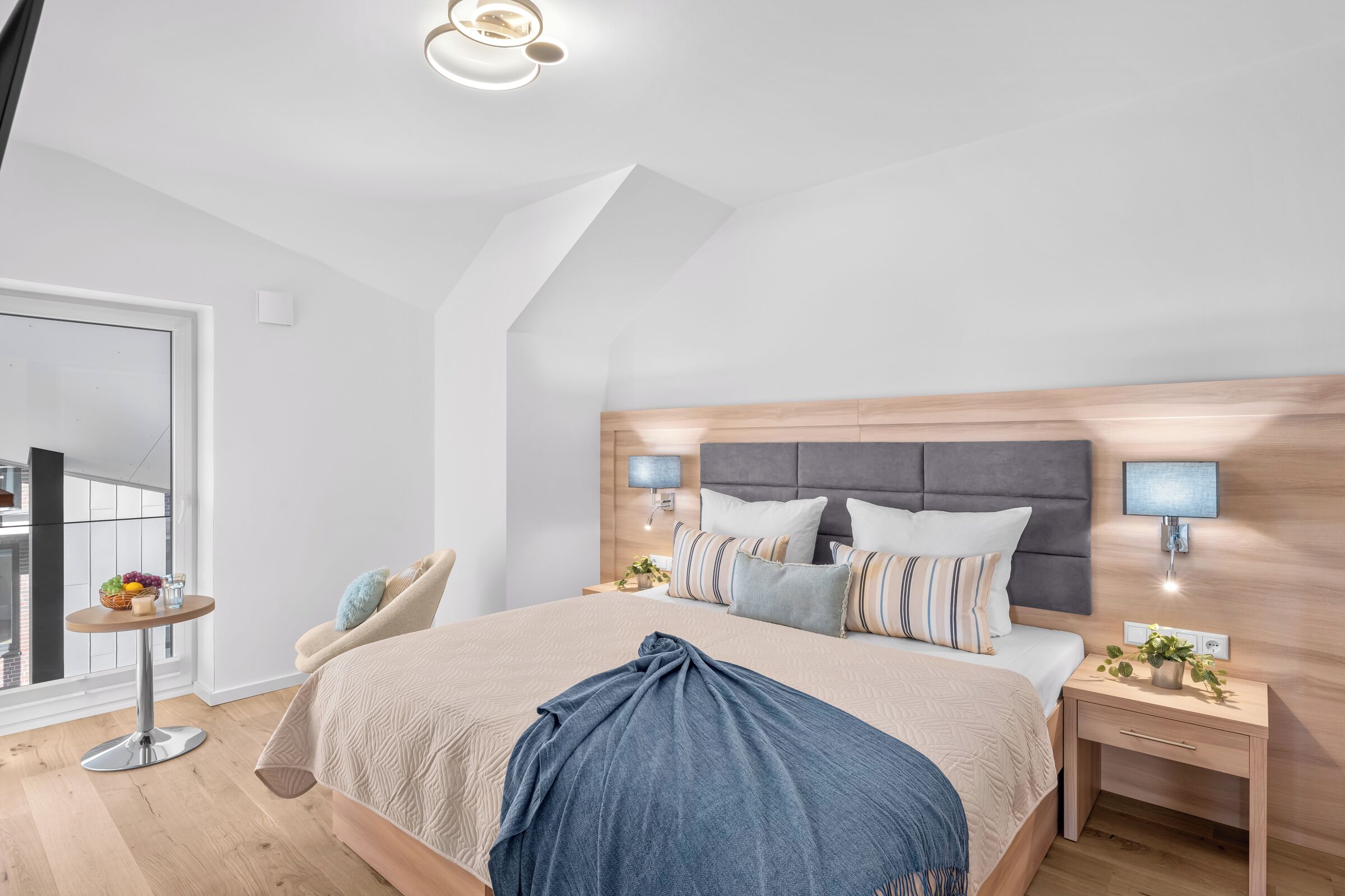 A modern bedroom with a large bed, bedside tables, a small table with fruit, and a view of the outside.