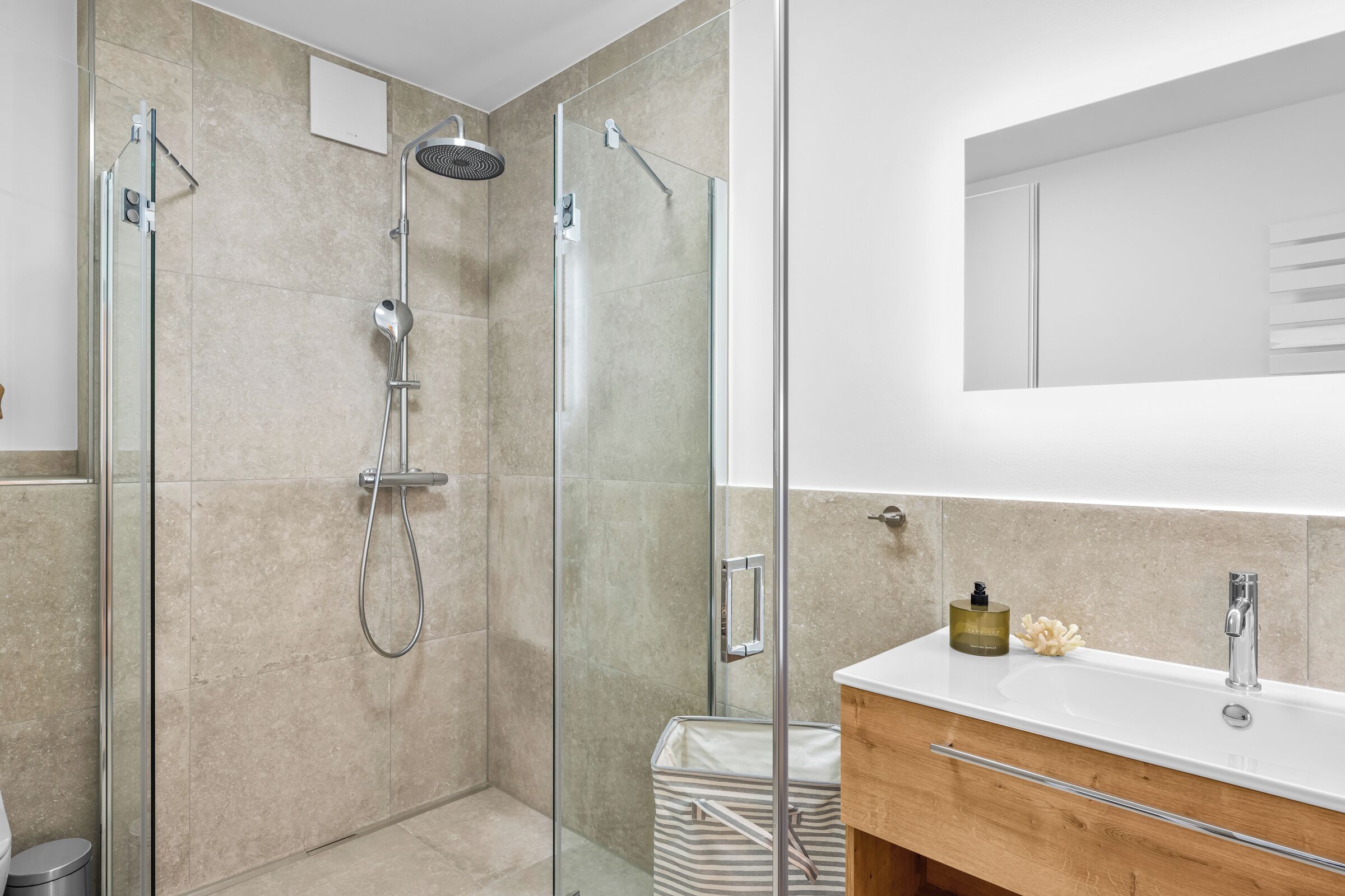 Een moderne badkamer met een glazen douchecabine, een witte wastafel en een houten opbergmeubel.