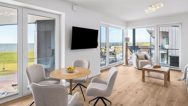 Ein modernes Wohnzimmer mit einem runden Holztisch, grauen Polstersesseln, einem flachbildfernseher an der Wand und großen Glasfenstertüren, die zu einem Außenbereich mit Meerblick führen.