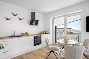 A modern kitchen with a built-in oven, a dining area with a round table and chairs, and a large window offering a view of the cityscape. - The View B14 Nordseeperle – Balcony, sea view, shared sauna (Dagebüll)