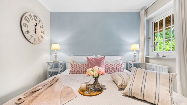 A bedroom with a bed, a clock, a window, and a bedside table with a lamp.