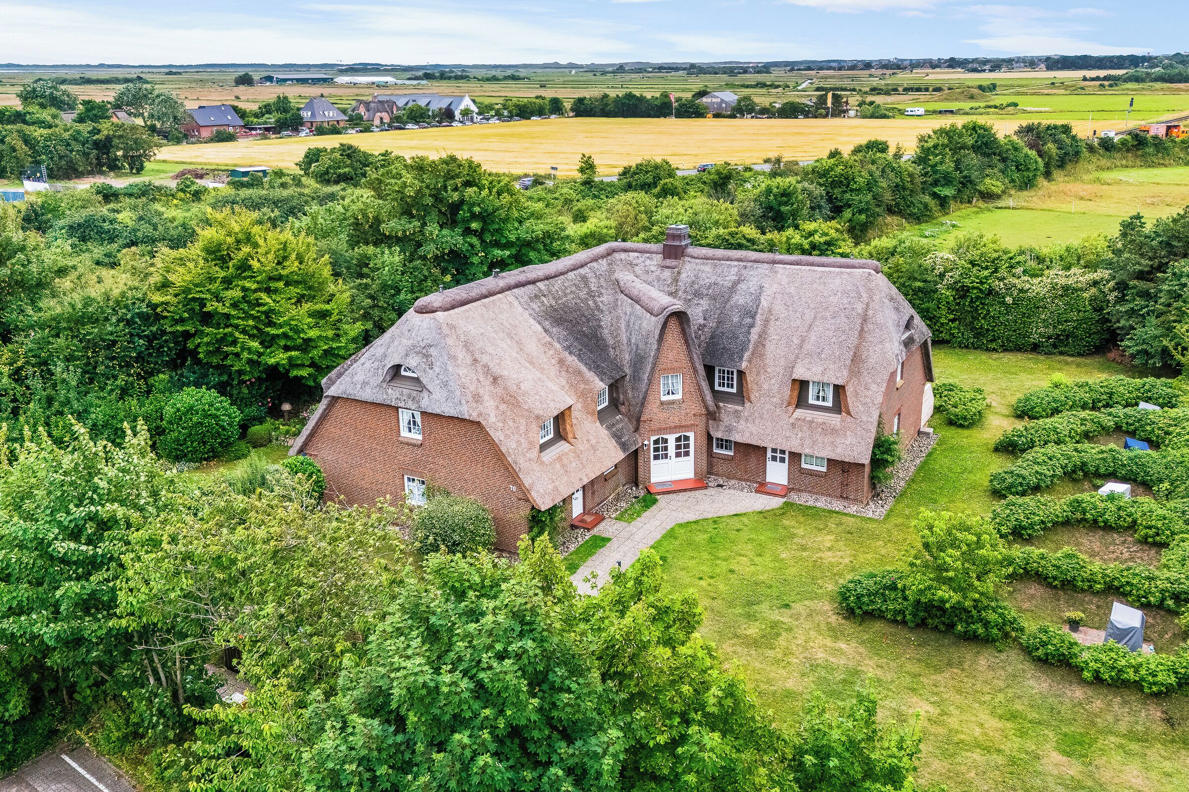 A large house with a steeply pitched roof, surrounded by lush greenery and fields.