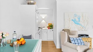 A modern kitchen with white cabinets, a dishwasher, and a sink. There is a dining area with a table set for two, a vase with flowers, and a bowl of fruit. A sofa and a coffee table are in the living area, and a painting hangs on the wall.