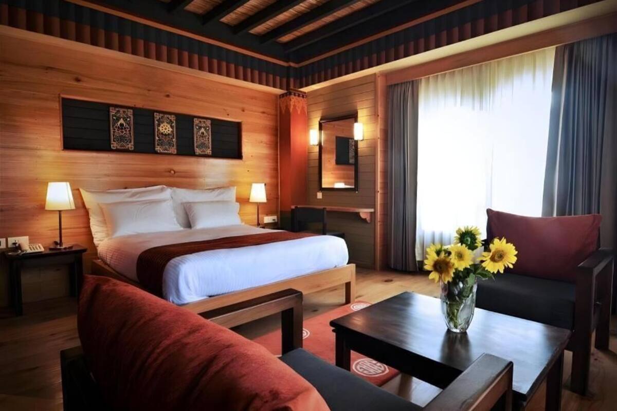 A wooden-furnished room with a bed, two bedside tables, a sofa, a coffee table, and a vase with flowers.