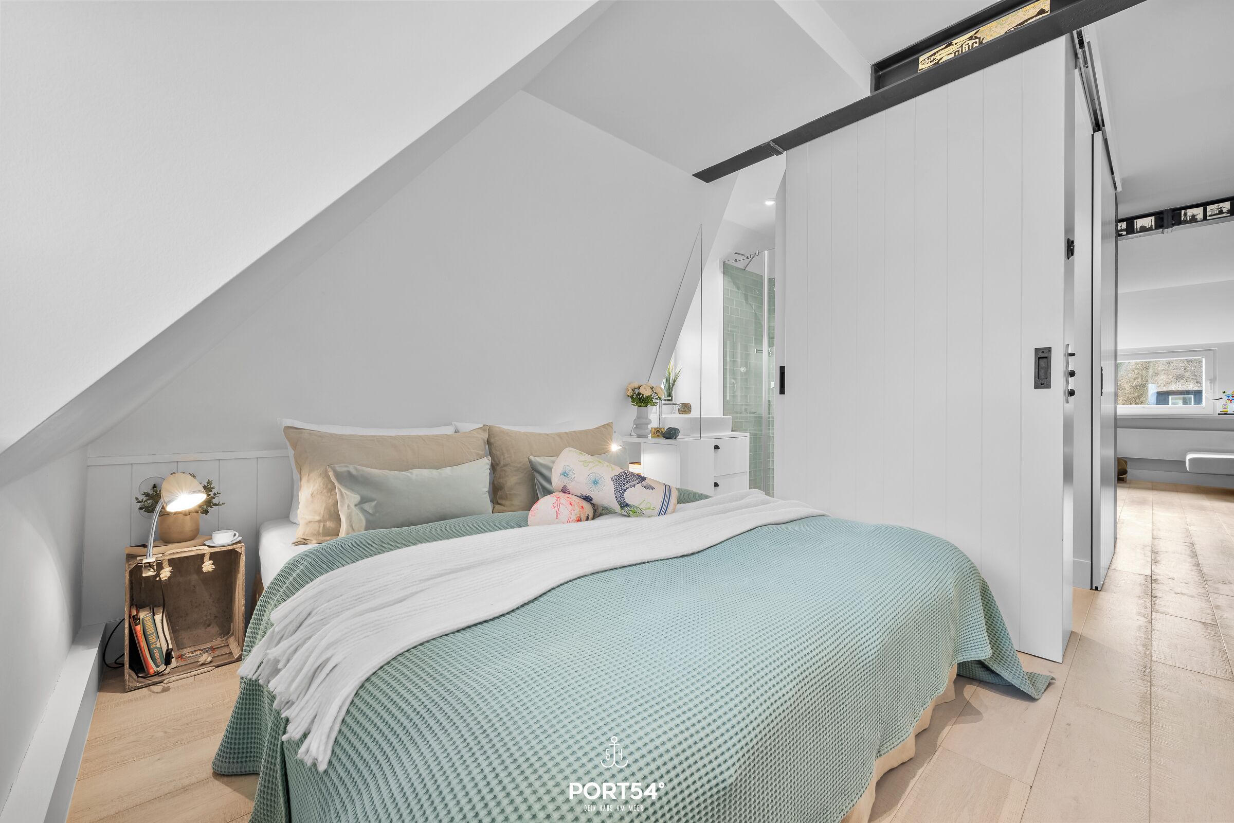 Ein Schlafzimmer mit schrägen Wänden, einem Bett mit grüner Bettdecke, einem Nachttisch mit Lampe und einer Kommode.