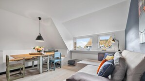 A loft-style room with a dining area, a sofa, and a window with a view of buildings. - High-quality vacation apartment in List on Sylt by the beach, with sauna and parking space (List auf Sylt)