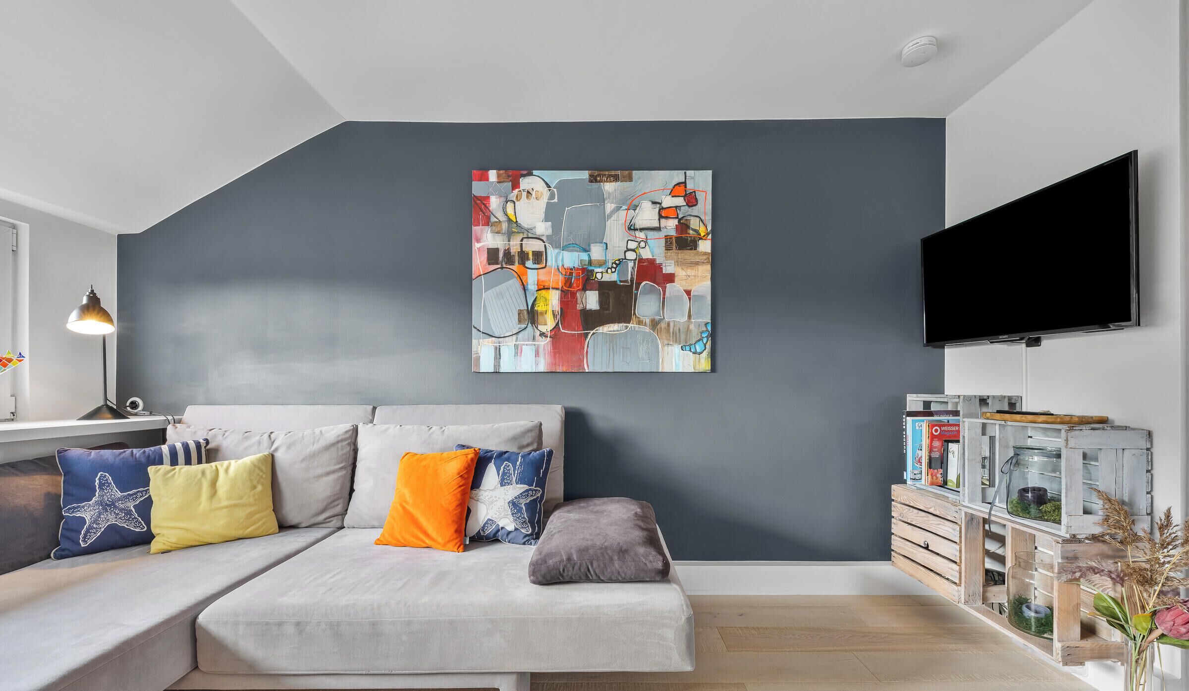 Een moderne woonkamer met een grijze bank, kleurrijke kussens, een groot abstract schilderij, een flatscreen televisie en een rustieke houten bijzettafel met een vaas.
