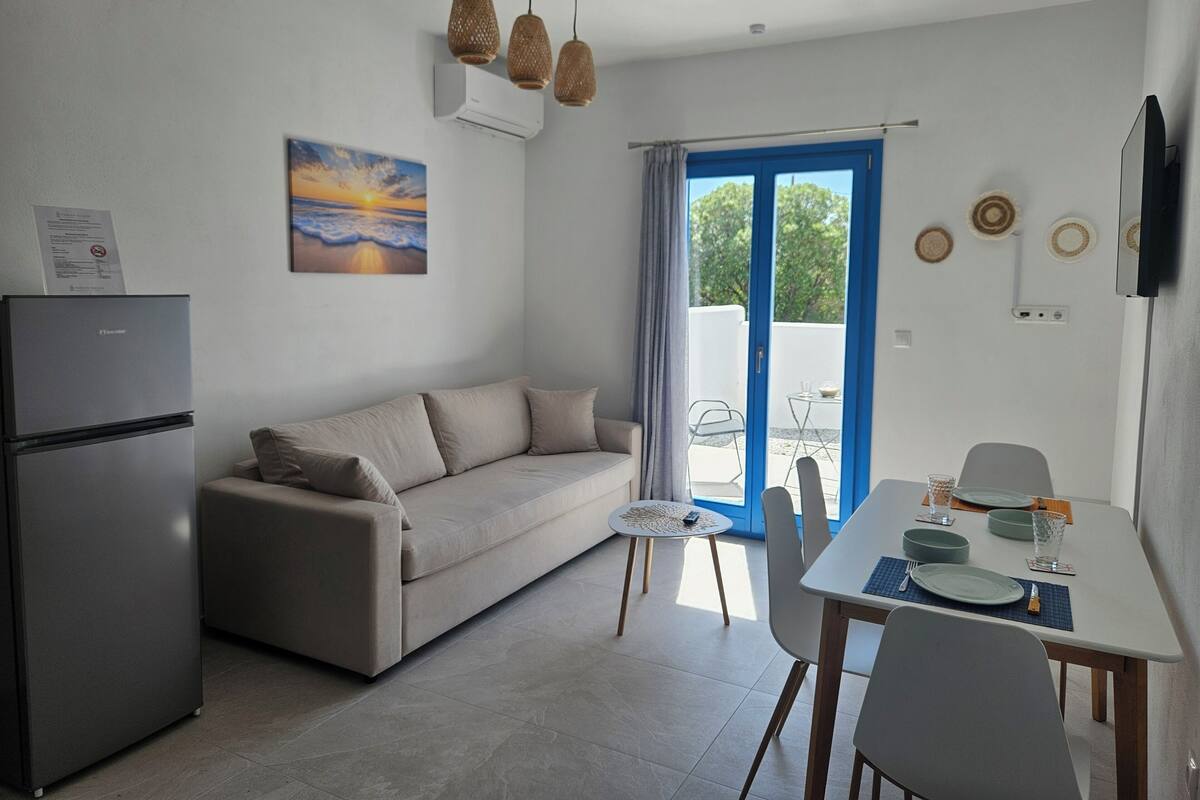 Un espace de vie moderne comprenant un canapé, une table à manger, des chaises, un réfrigérateur et une vue sur l’extérieur, visible à travers une porte bleue.