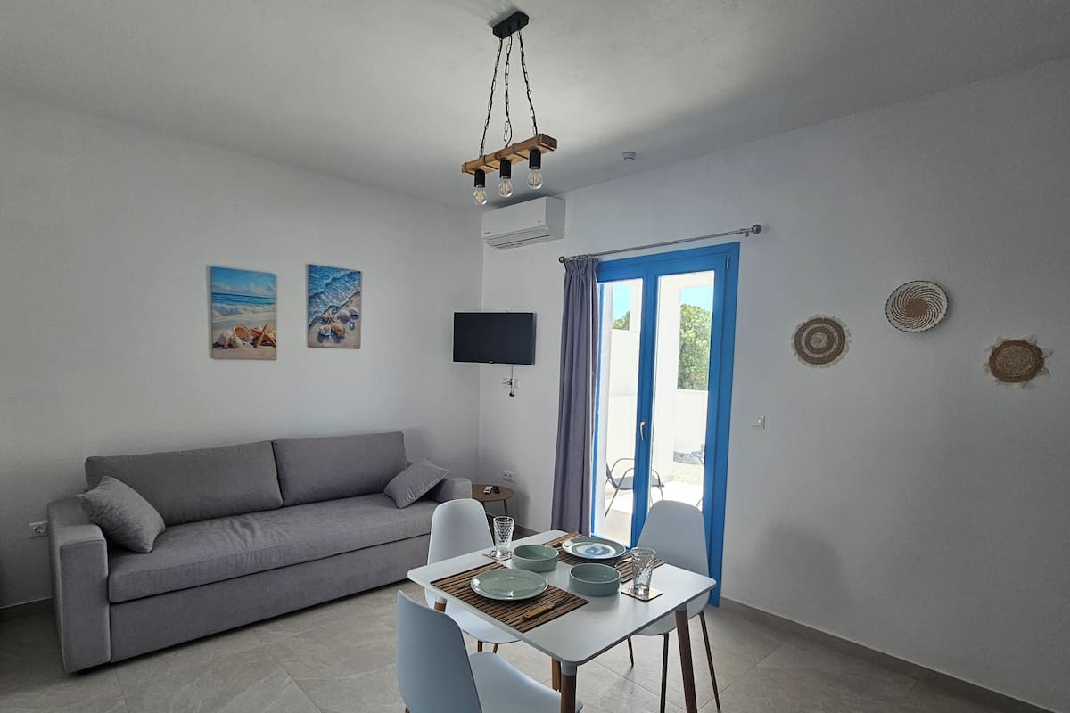Un salon moderne avec un canapé gris, une table à manger avec des chaises, un téléviseur à écran plat et un luminaire suspendu au plafond.