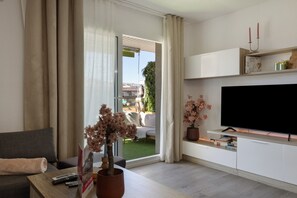 Un soggiorno moderno con un divano, un tavolino da caffè con telecomandi e un libro, una TV su un mobile e una porta vetrata scorrevole che dà su uno spazio esterno.