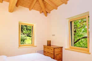 A bedroom with a bed, wooden dresser, two windows with views of greenery, and a wooden ceiling.