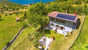 Une maison dotée d’un toit recouvert de panneaux solaires, entourée de verdure et d’un mur de pierres, offrant une vue sur un lac et des montagnes en arrière-plan.